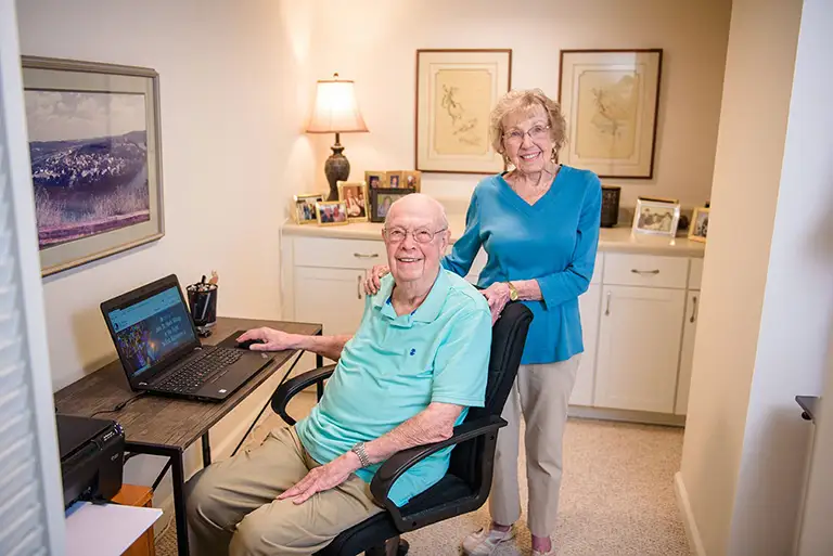 An elderly man sits at a desk with a laptop while an elderly woman stands beside him, both smiling, in a St. Mark Village senior living home office adorned with family photos and framed artwork. An elderly man sits at a desk with a laptop while an elderly woman stands beside him, both smiling, in a St. Mark Village senior living home office adorned with family photos and framed artwork.