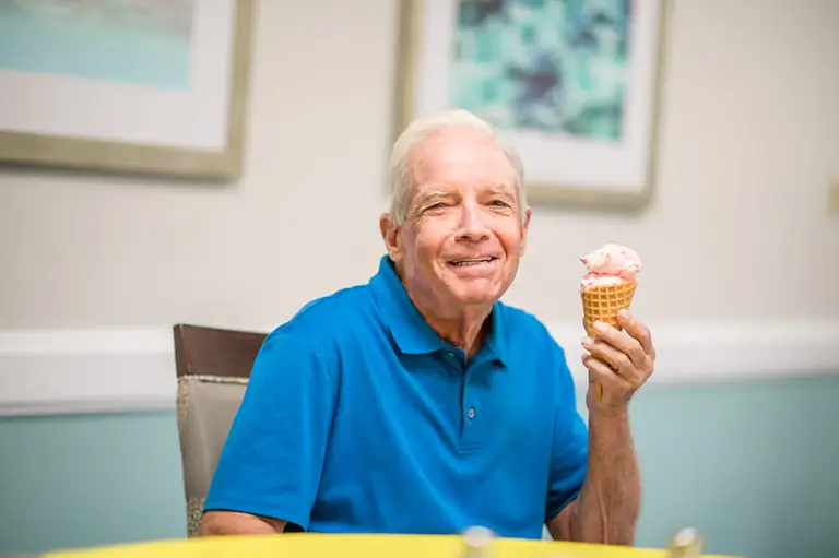 Older man in a blue polo shirt sits at a table, smiling and holding an ice cream cone—enjoying the sweet moments of senior living at St. Mark Village. Older man in a blue polo shirt sits at a table, smiling and holding an ice cream cone—enjoying the sweet moments of senior living at St. Mark Village.