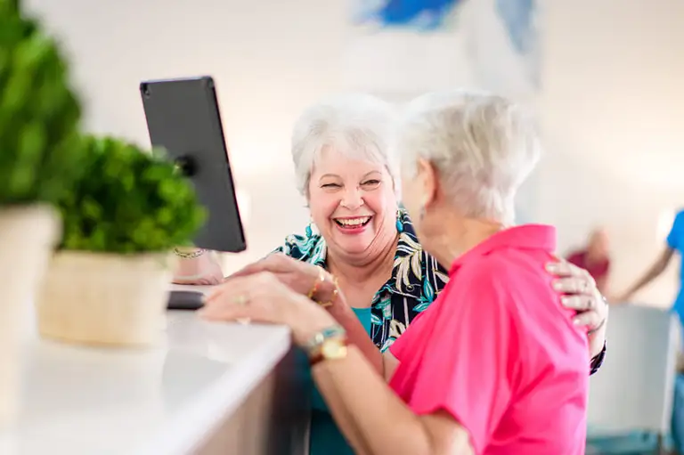 Two older women interact at a counter in St. Mark Village senior living, one smiling broadly while the other has her back to the camera. A tablet and potted plant sit on the counter. Two older women interact at a counter in St. Mark Village senior living, one smiling broadly while the other has her back to the camera. A tablet and potted plant sit on the counter.