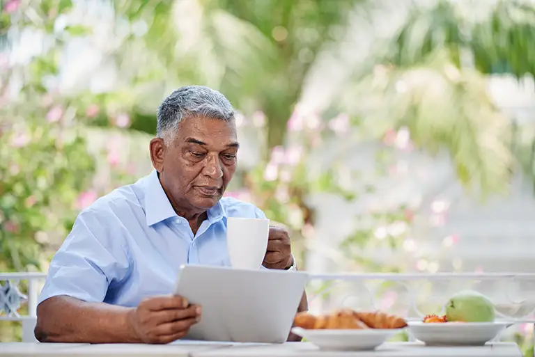 An older man sits at an outdoor table in St. Mark Village senior living, holding a white mug and looking at a tablet. Plates of food and fruit are on the table, with lush greenery in the background. An older man sits at an outdoor table in St. Mark Village senior living, holding a white mug and looking at a tablet. Plates of food and fruit are on the table, with lush greenery in the background.