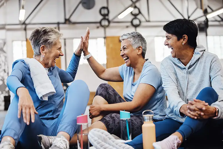 Three older adults in athletic wear sit on a gym floor at St. Mark Village, smiling and giving high-fives, with water bottles nearby—celebrating an active approach to senior living. Three older adults in athletic wear sit on a gym floor at St. Mark Village, smiling and giving high-fives, with water bottles nearby—celebrating an active approach to senior living.