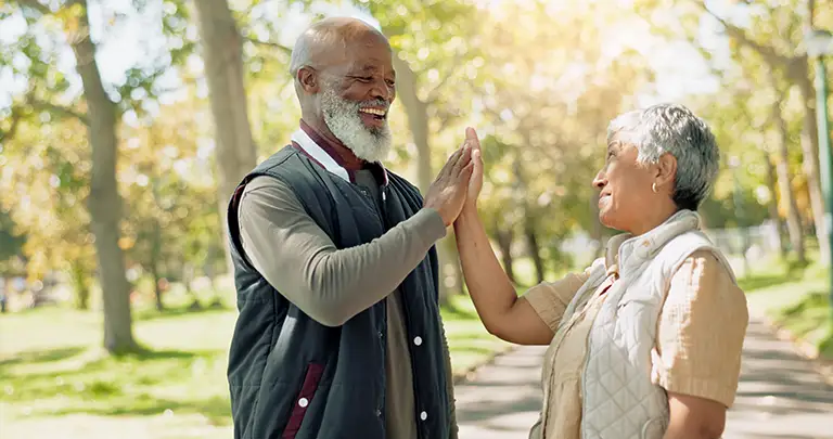 Two older adults outside in a park at St. Mark Village, smiling and giving each other a high five on a sunny day, enjoying vibrant senior living. Two older adults outside in a park at St. Mark Village, smiling and giving each other a high five on a sunny day, enjoying vibrant senior living.