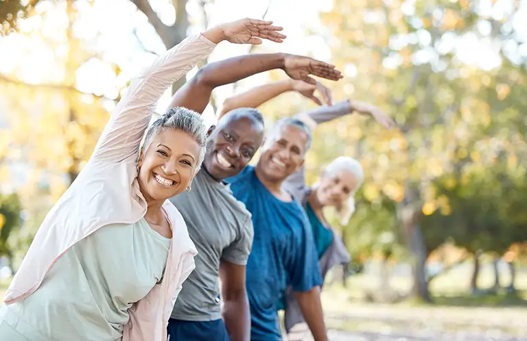 Four adults stand outdoors in a row at St. Mark Village, smiling and stretching with one arm overhead. Trees and sunlight brighten the background, reflecting the vibrant senior living lifestyle. Four adults stand outdoors in a row at St. Mark Village, smiling and stretching with one arm overhead. Trees and sunlight brighten the background, reflecting the vibrant senior living lifestyle.