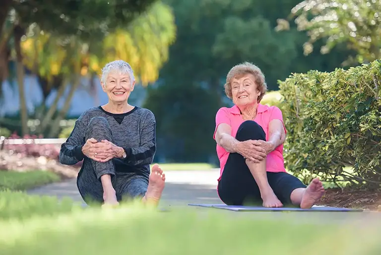 Two older women sit on the ground outdoors at St. Mark Village, smiling with legs bent and arms wrapped around their knees, surrounded by lush greenery—a perfect moment of connection in senior living. Two older women sit on the ground outdoors at St. Mark Village, smiling with legs bent and arms wrapped around their knees, surrounded by lush greenery—a perfect moment of connection in senior living.