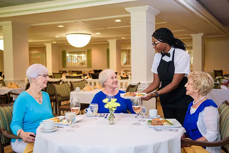 A server in uniform delivers plates of food to three elderly women seated at a round table in the bright, welcoming dining room of St. Mark Village, highlighting the warm community atmosphere of senior living.