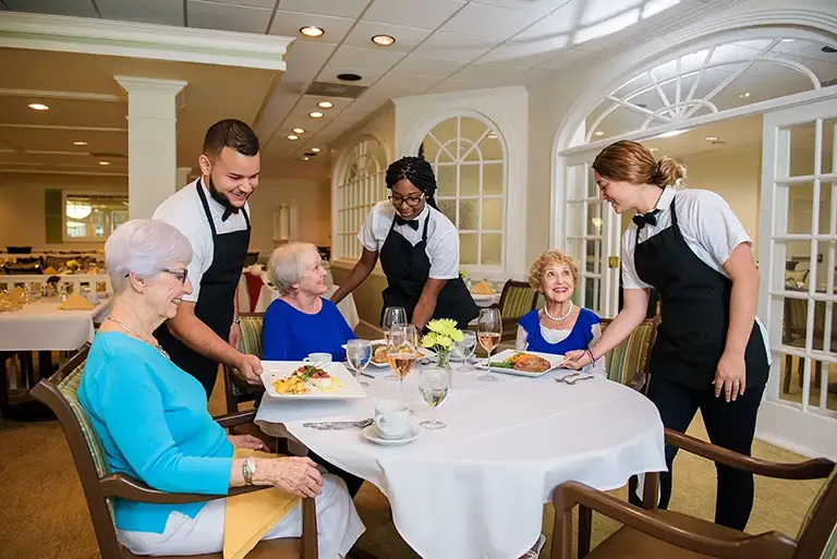 Three servers deliver meals to three elderly women seated at a round dining table in the well-lit, upscale dining room at St. Mark Village, a premier senior living community. Three servers deliver meals to three elderly women seated at a round dining table in the well-lit, upscale dining room at St. Mark Village, a premier senior living community.