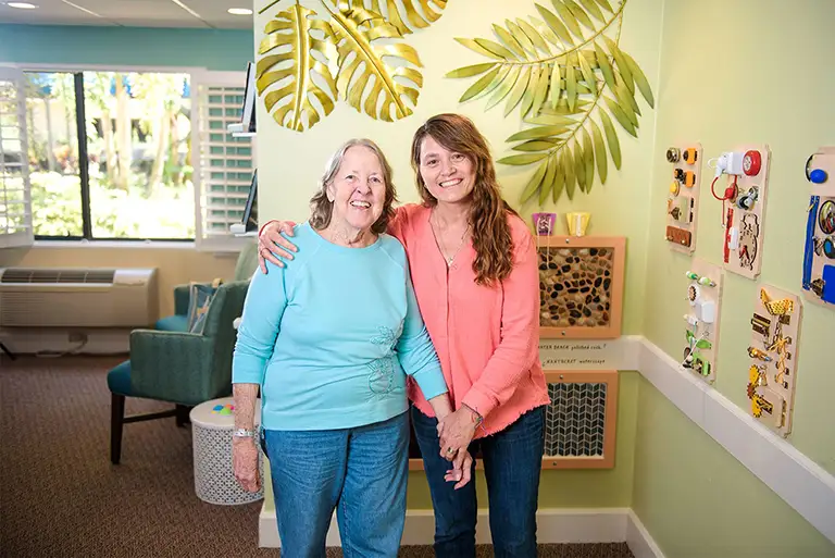 Two women stand together in a brightly lit room at St. Mark Village, with large leaf decorations on the wall, smiling at the camera and showcasing the vibrant spirit of senior living.