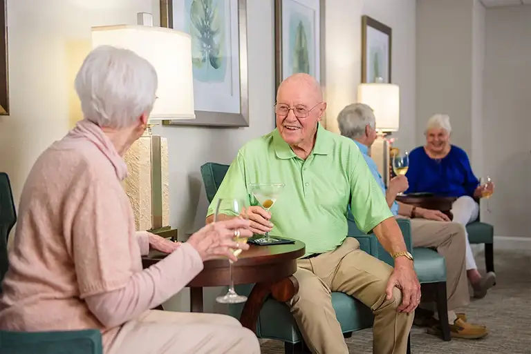 Four older adults sit in a lounge area at St. Mark Village, holding drinks and conversing. The relaxed, social senior living setting features framed art and lamps in the background. Four older adults sit in a lounge area at St. Mark Village, holding drinks and conversing. The relaxed, social senior living setting features framed art and lamps in the background.