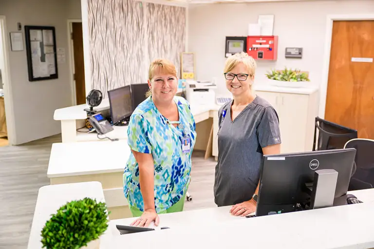 Two women in medical scrubs stand and smile at a reception desk in St. Mark Village, a senior living healthcare facility, with office equipment and plants visible in the background. Two women in medical scrubs stand and smile at a reception desk in St. Mark Village, a senior living healthcare facility, with office equipment and plants visible in the background.
