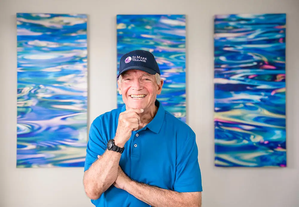Smiling older man in a blue shirt and cap stands in front of three abstract blue water-themed paintings at St. Mark Village, reflecting the vibrant spirit of senior living.