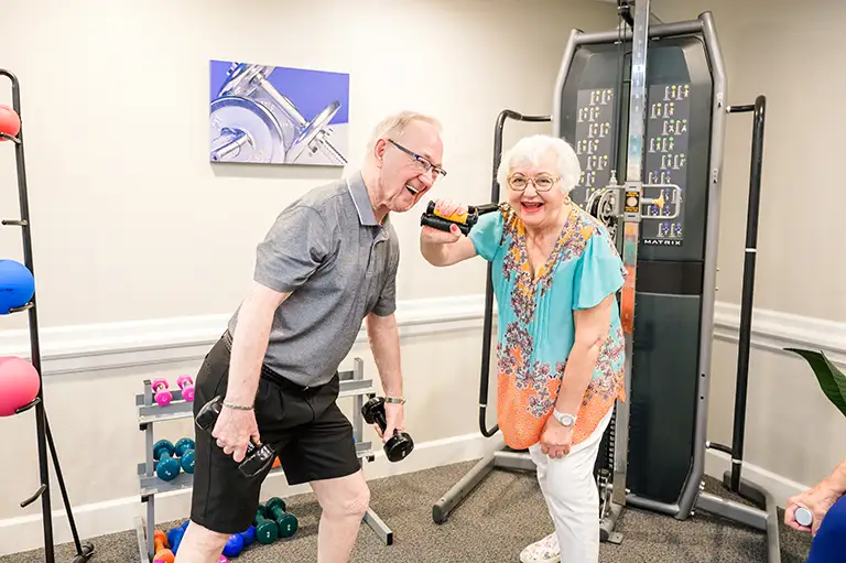 Two older adults exercise with dumbbells in a gym at St. Mark Village senior living, smiling near weight racks and a weight machine. Two older adults exercise with dumbbells in a gym at St. Mark Village senior living, smiling near weight racks and a weight machine.