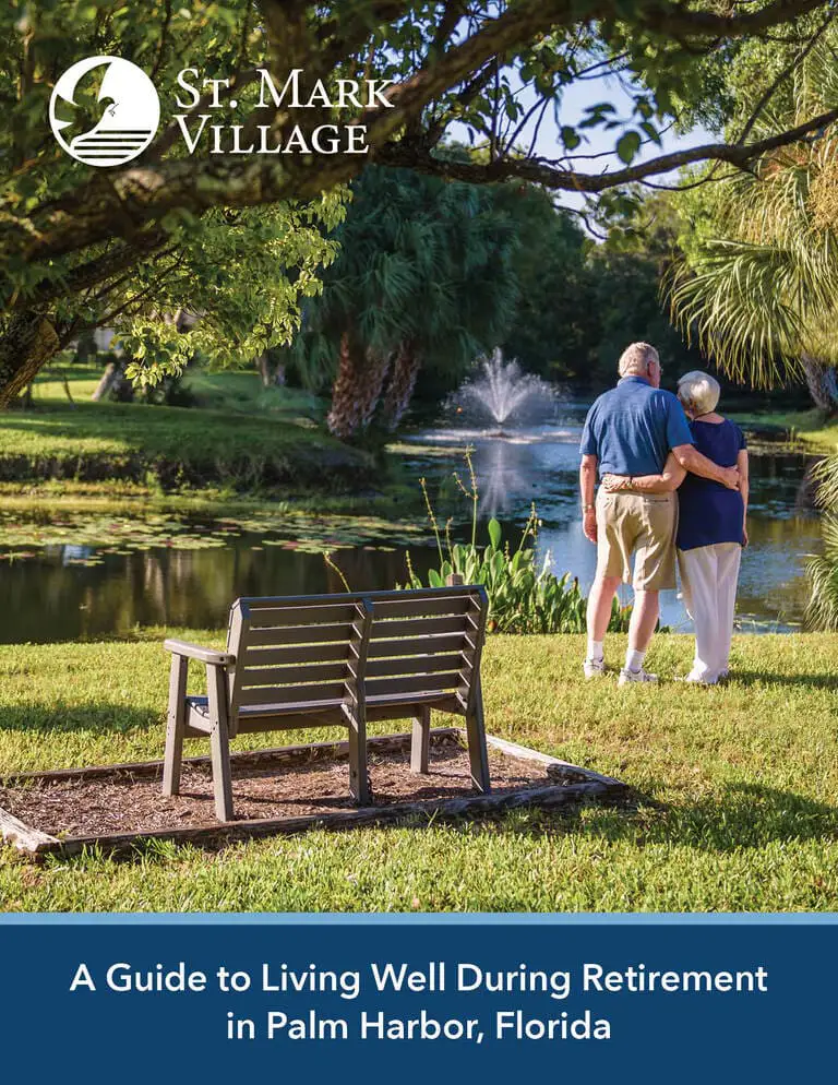 An elderly couple stands by a pond with a fountain, embracing near a bench in the lush green park at St. Mark Village senior living, Palm Harbor, Florida.