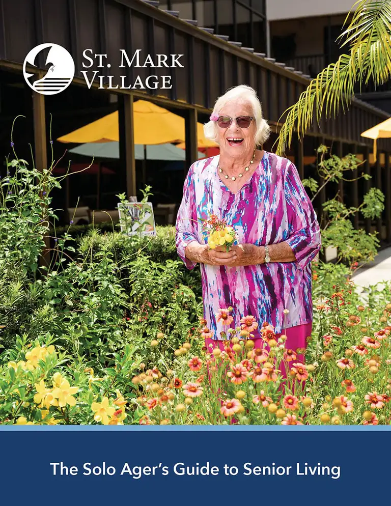 An older woman in a colorful dress stands smiling in a garden outside a building with the text "St. Mark Village," showcasing the joys of senior living in "The Solo Ager’s Guide to Senior Living.