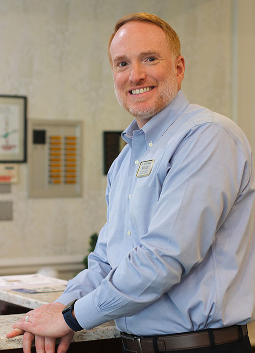 A man with short red hair and a beard, wearing a light blue dress shirt and name tag, stands smiling at a counter in the St. Mark Village senior living reception area. A man with short red hair and a beard, wearing a light blue dress shirt and name tag, stands smiling at a counter in the St. Mark Village senior living reception area.