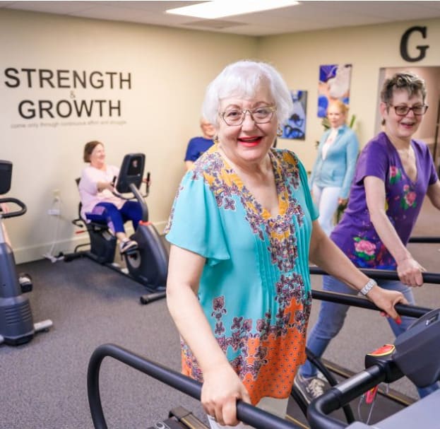 Elderly women in St. Mark Village's senior living community exercise on treadmills and stationary bikes in a fitness room with "Strength & Growth" written on the wall.