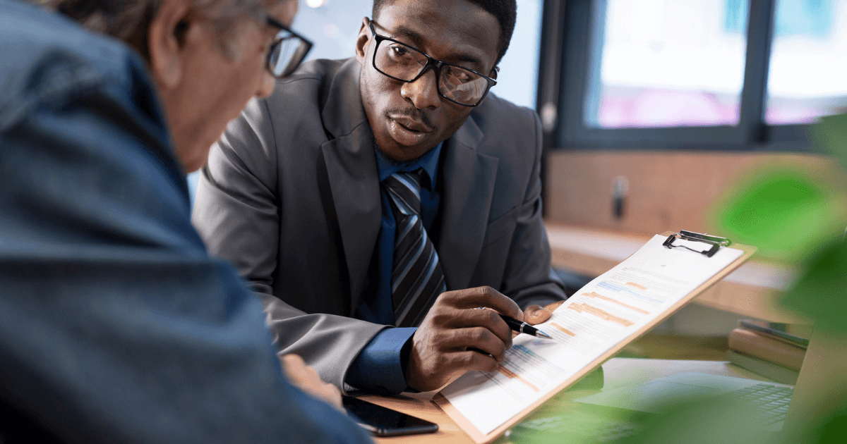 A man in a suit shows and discusses a document on a clipboard with another person across a desk, highlighting senior living options at St. Mark Village in an office setting.