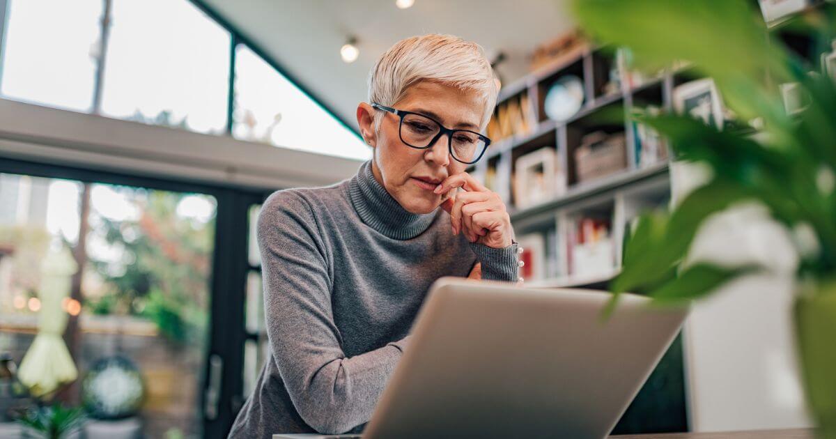 A person with short gray hair and glasses, wearing a gray turtleneck, sits at a table working on a laptop in a modern, bright home office at St. Mark Village, embracing the comforts of senior living.