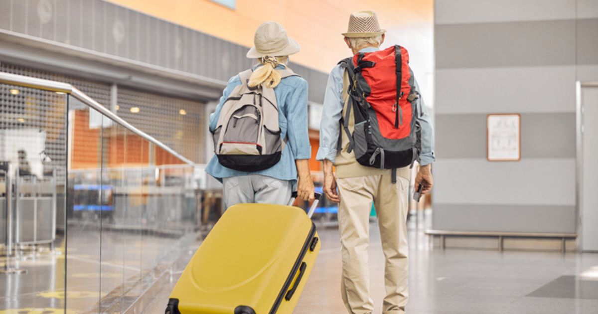 Two older adults wearing hats walk through an airport terminal, each with a backpack and one pulling a yellow suitcase—embracing the spirit of senior living at St. Mark Village.