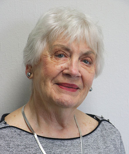 An older woman with short white hair and a light gray top, wearing a silver necklace and earrings, stands in front of a plain, light-colored wall at St. Mark Village senior living community. An older woman with short white hair and a light gray top, wearing a silver necklace and earrings, stands in front of a plain, light-colored wall at St. Mark Village senior living community.