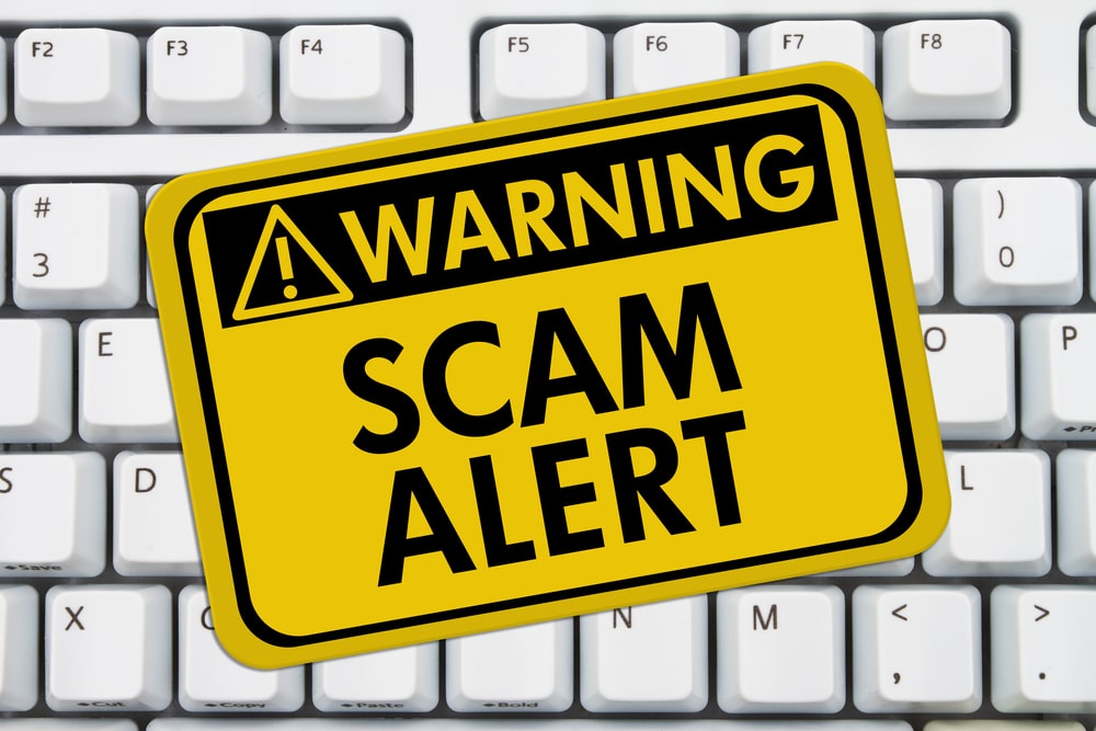 A yellow "Warning Scam Alert" sign is placed on top of a white computer keyboard, highlighting online security for seniors at St. Mark Village senior living community.