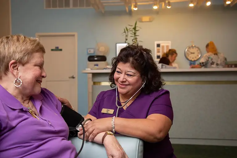 A nurse at St. Mark Village checks a patient’s blood pressure in a medical clinic, both women smiling and wearing purple uniforms; two staff members work at the reception desk in the background, highlighting quality senior living care. A nurse at St. Mark Village checks a patient's blood pressure in a medical clinic, both women smiling and wearing purple uniforms; two staff members work at the reception desk in the background, highlighting quality senior living care.