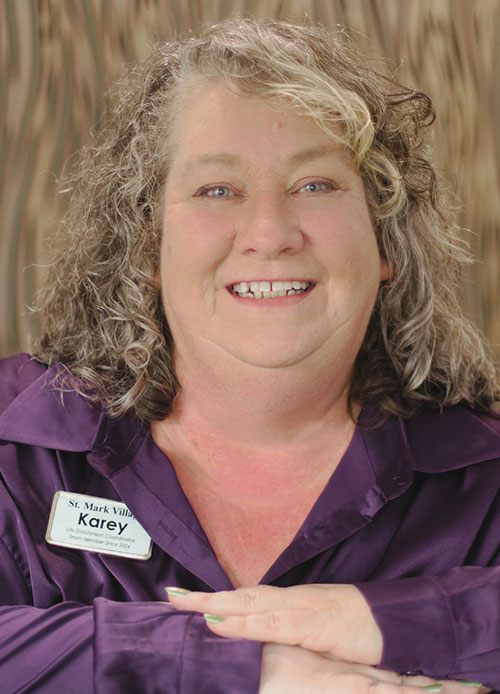 A woman with curly gray hair wearing a purple blouse and a St. Mark Village name tag that reads “Karey” smiles at the camera with her arms crossed in front of her, reflecting the warmth of senior living. A woman with curly gray hair wearing a purple blouse and a St. Mark Village name tag that reads “Karey” smiles at the camera with her arms crossed in front of her, reflecting the warmth of senior living.