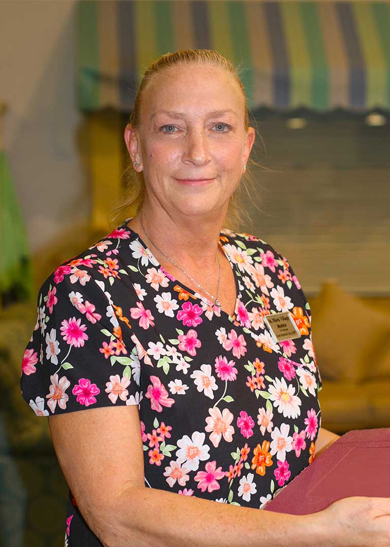 A woman with blonde hair in a ponytail, wearing a floral scrub top and a name badge, stands indoors at St. Mark Village holding a maroon folder, smiling at the camera—a warm presence in senior living. A woman with blonde hair in a ponytail, wearing a floral scrub top and a name badge, stands indoors at St. Mark Village holding a maroon folder, smiling at the camera—a warm presence in senior living.