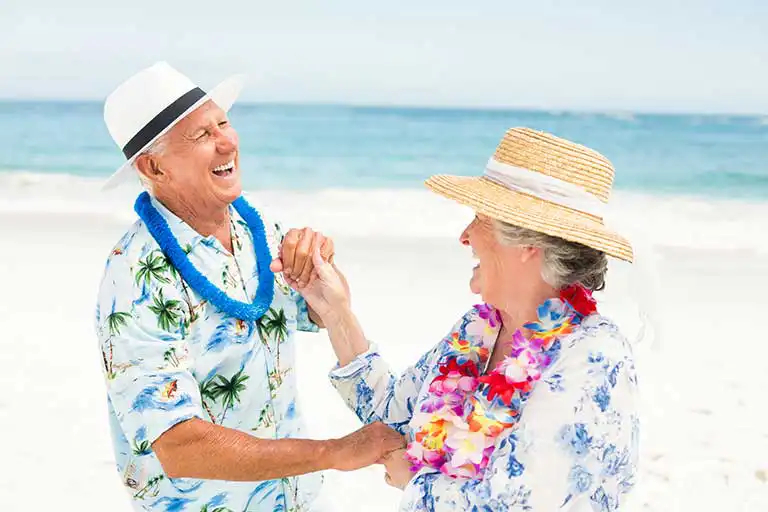 Two older adults wearing leis and sun hats laugh and hold hands on a sunny beach, with the ocean and sky in the background—capturing the vibrant spirit of senior living at St. Mark Village. Two older adults wearing leis and sun hats laugh and hold hands on a sunny beach, with the ocean and sky in the background—capturing the vibrant spirit of senior living at St. Mark Village.
