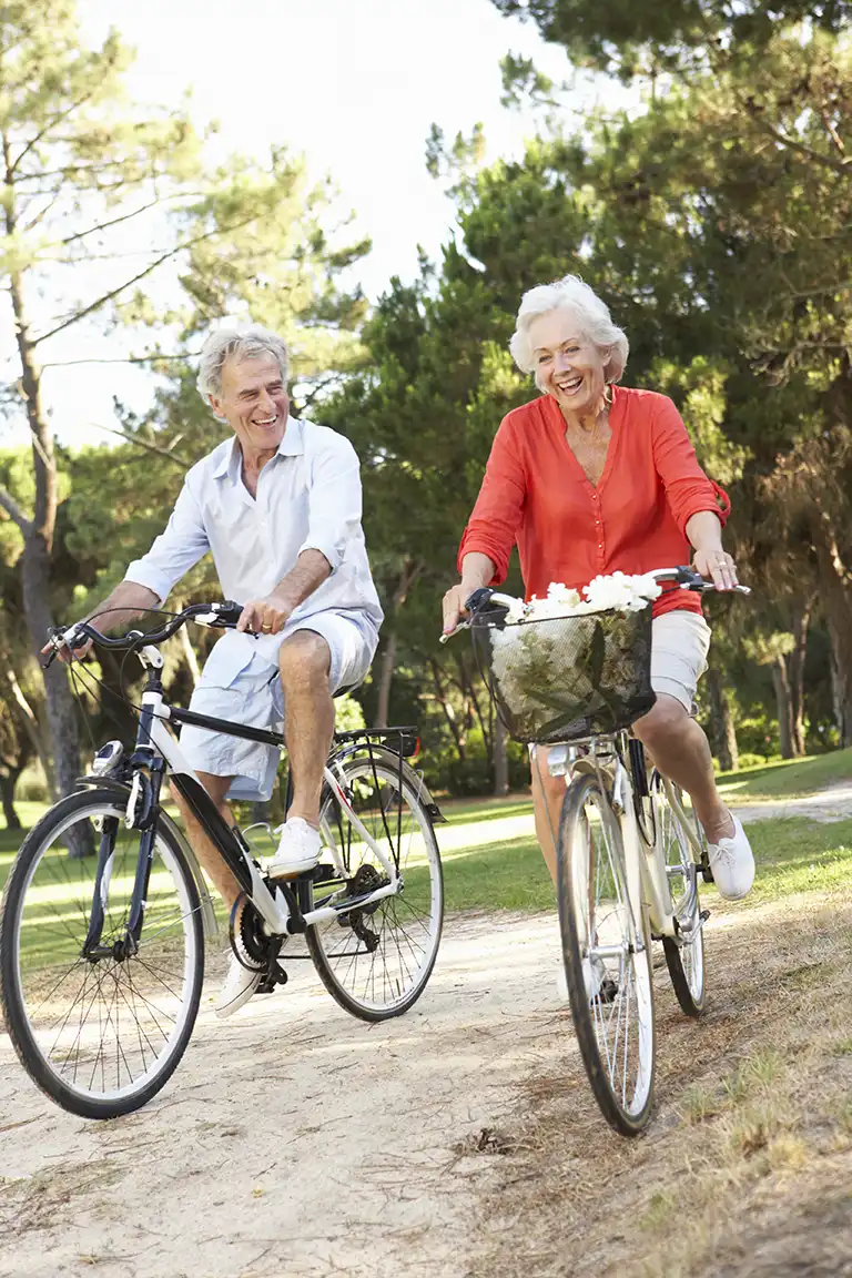 Residents of St. Mark Village enjoy senior living as an older man and woman ride bicycles on a dirt path through a park, surrounded by trees. The woman has a basket of flowers on her bike. Residents of St. Mark Village enjoy senior living as an older man and woman ride bicycles on a dirt path through a park, surrounded by trees. The woman has a basket of flowers on her bike.