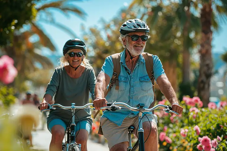 Two older adults wearing helmets and sunglasses ride bicycles on a sunny day, surrounded by palm trees and blooming flowers, enjoying the vibrant lifestyle of senior living at St. Mark Village.