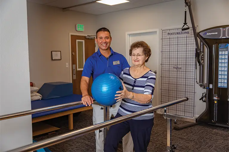 A physical therapist assists an older woman holding an exercise ball while standing between parallel bars in the St. Mark Village senior living rehabilitation clinic.