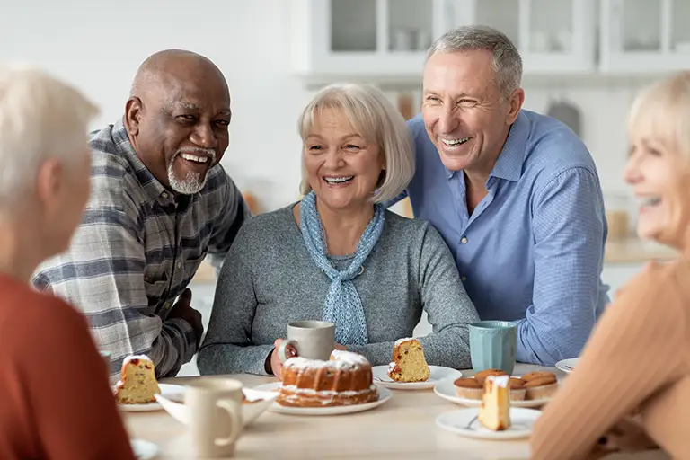 A group of older adults sits around a table with coffee and cake, smiling and chatting in a bright kitchen at St. Mark Village, enjoying the warmth of community and vibrant senior living.