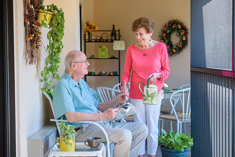An older man sits on a porch at St. Mark Village, reading a newspaper while an older woman stands nearby with a watering can, surrounded by potted plants and patio furniture—a peaceful scene of senior living. An older man sits on a porch at St. Mark Village, reading a newspaper while an older woman stands nearby with a watering can, surrounded by potted plants and patio furniture—a peaceful scene of senior living.