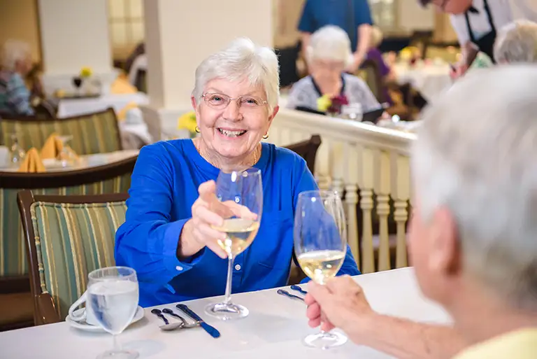 An older woman in a blue shirt smiles and raises a wine glass in a toast at a dining table with another person, enjoying the vibrant senior living community at St. Mark Village. An older woman in a blue shirt smiles and raises a wine glass in a toast at a dining table with another person, enjoying the vibrant senior living community at St. Mark Village.