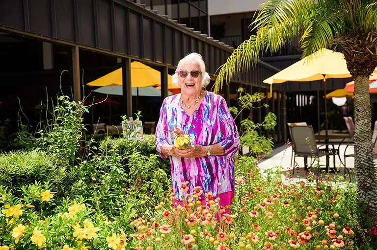 An elderly woman in a colorful blouse stands in a garden with blooming flowers at St. Mark Village senior living, holding a plant. Patio umbrellas and outdoor seating are visible in the background.